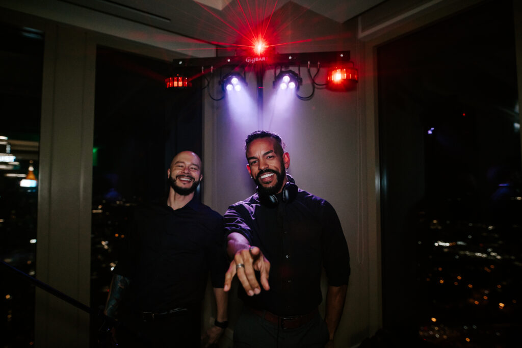Two people dressed in black are standing under red and purple lights. One is pointing towards the camera. Both appear to be in a dark room with city lights visible through the windows in the background.