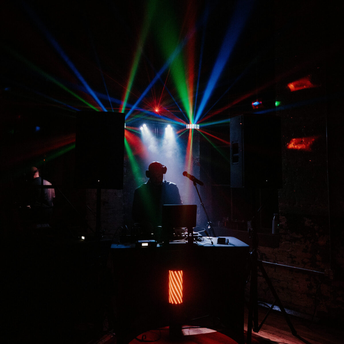 A DJ performs on stage with colorful laser lights and fog effects, surrounded by speakers and music equipment.